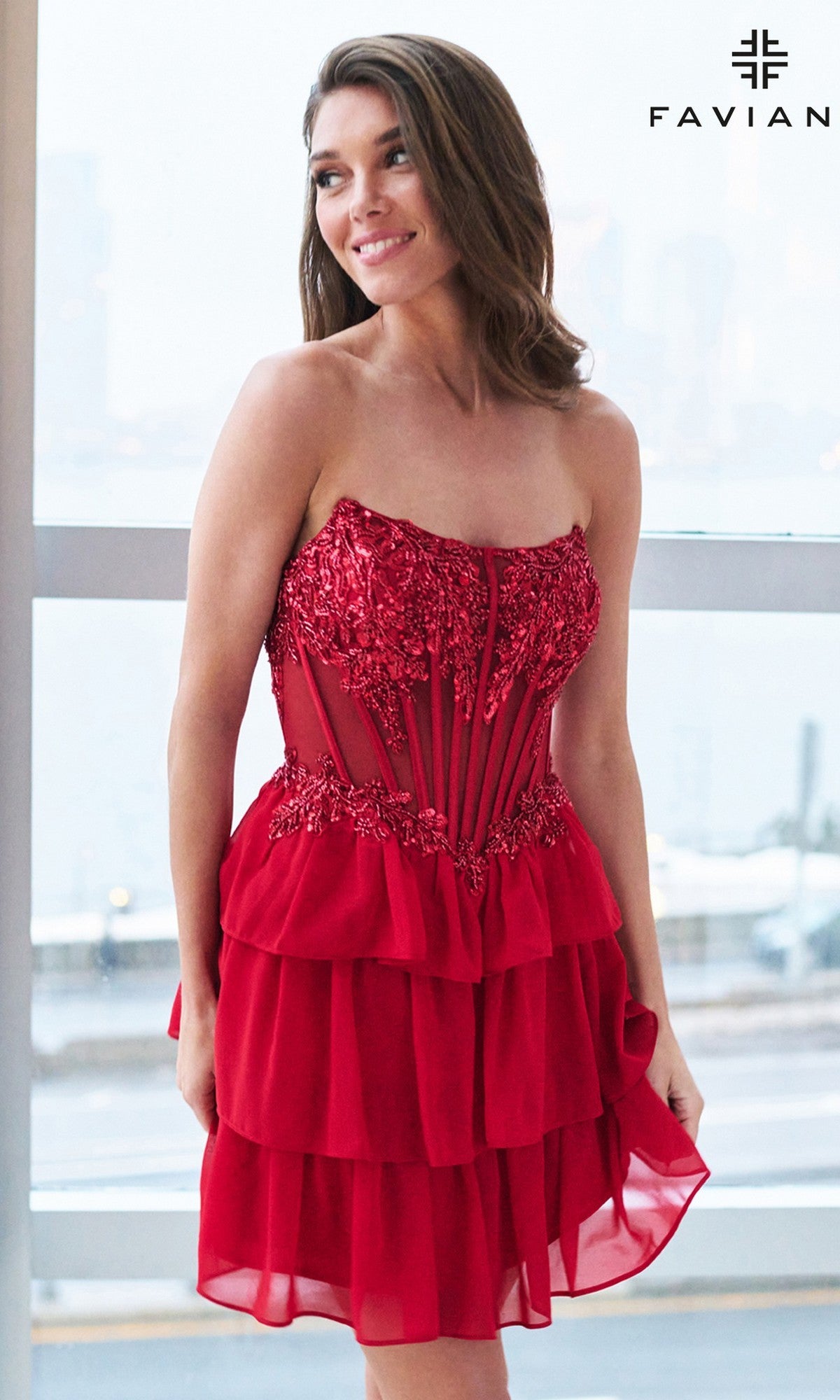 A woman wearing the Faviana 11500 short homecoming dress with a strapless corset bodice, lace, and sheer detailing stands smiling by a window indoors. The